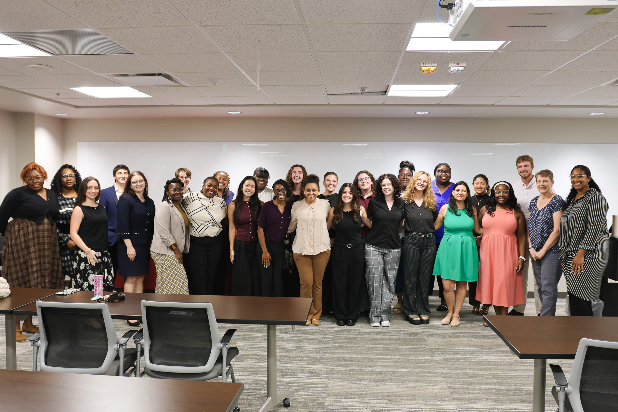 Large group of students and faculty posing together in a classroom with tables, chairs, and a whiteboard.