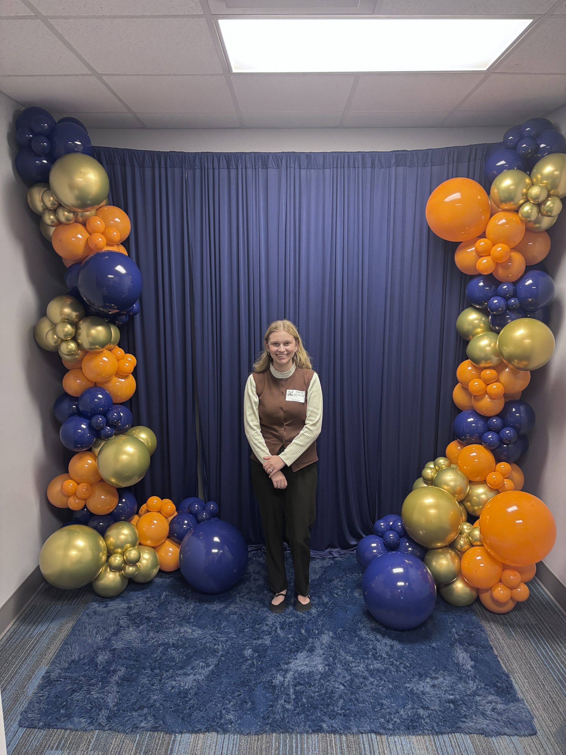 Student stands in front of a blue curtain backdrop framed by blue, gold, and orange balloon displays.
