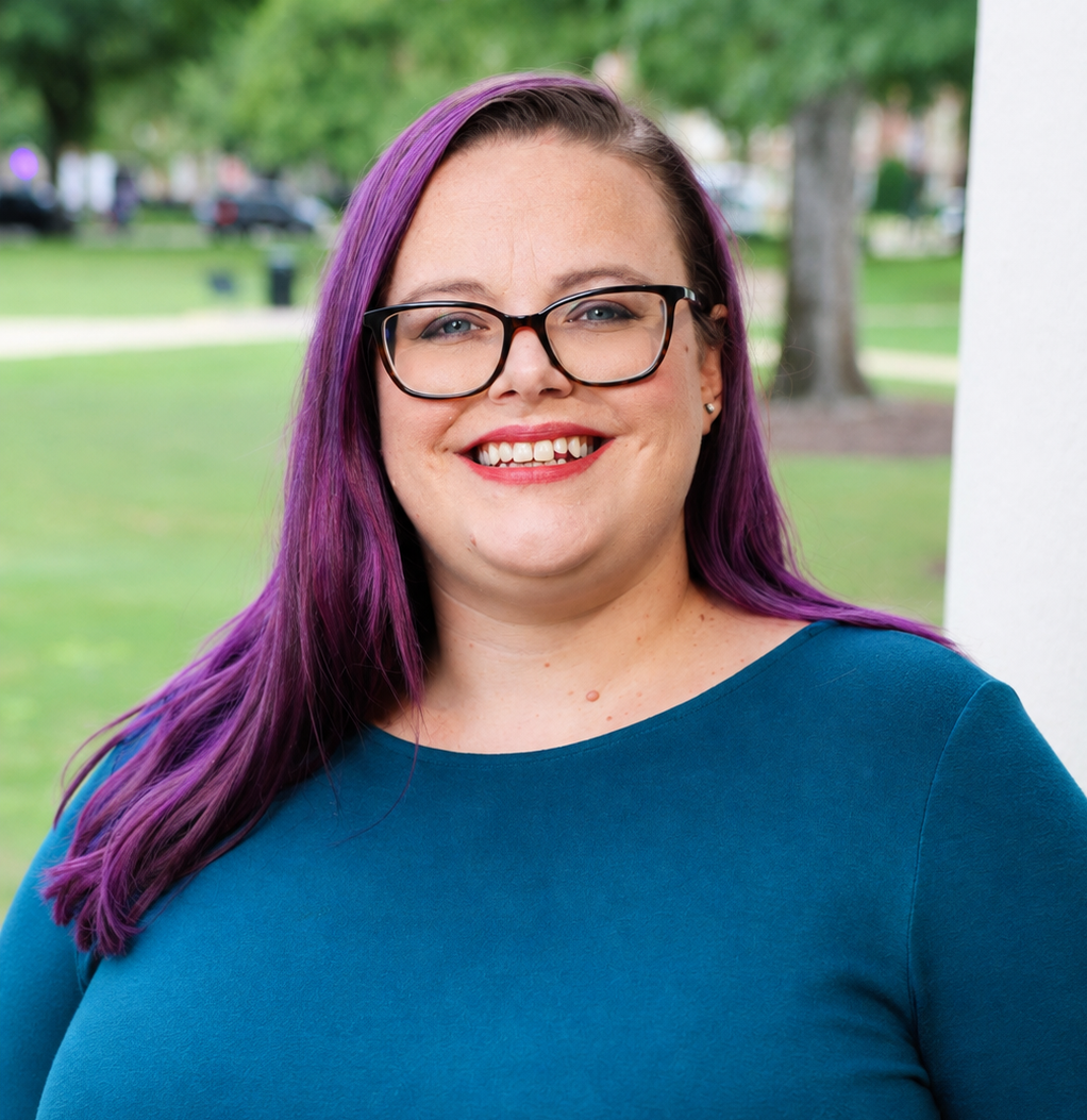 Person with purple hair and glasses standing outdoors on a campus lawn, wearing a blue top.